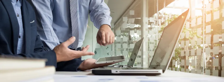 imagem mostra um computador sobre a mesa e é possível ver parte do corpo de duas pessoas conversando