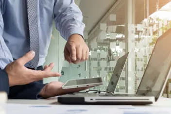 imagem mostra um computador sobre a mesa e é possível ver parte do corpo de duas pessoas conversando