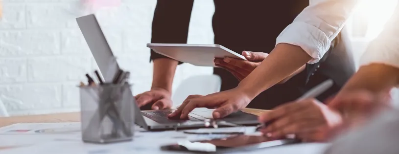 uma mesa com papeis e computador em que é possível ver as mãos de duas pessoas