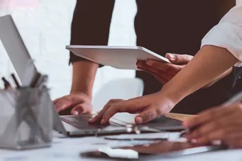 uma mesa com papeis e computador em que é possível ver as mãos de duas pessoas